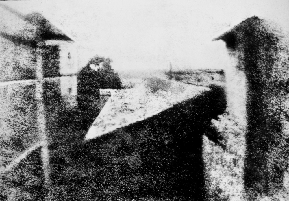 The world's first photograph taken by Joseph Nicéphore Niépce in 1826 or 1827. Captured using a technique known as heliography from an upstairs window at Niépce's estate in Burgundy. The image shows a granular black and white view of a small lawn stretching between two buildings. On the left building two small windows are visible. In the distance is a bush or several trees.