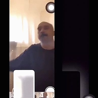 The right side of image is black, the left side of image shows a bald caucasian man with moustache in a black shirt. The image is blurry, as if taken with an old phone camera. The image is candid, the man seems relaxed and at home. There is a white dot above the man's head and a white square under his arm.